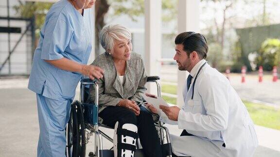 图为一名白人男性医生正在与腿上绑着夹板坐在轮椅上接受身体检查的亚洲老年妇女交谈