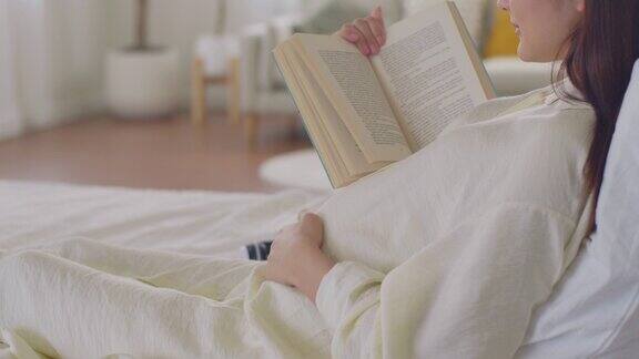 快乐的亚洲孕妇在家里的床上读书轻轻抚摸她的肚子