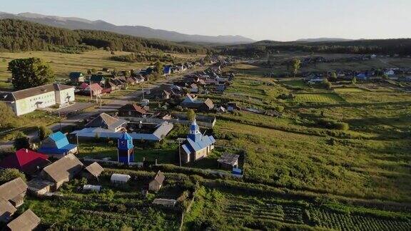 夏天俄罗斯村庄的鸟瞰图飞过木制房屋茂密的绿色植物和俄罗斯小定居点的田野太阳照亮了远处的群山和茂密的绿色森林