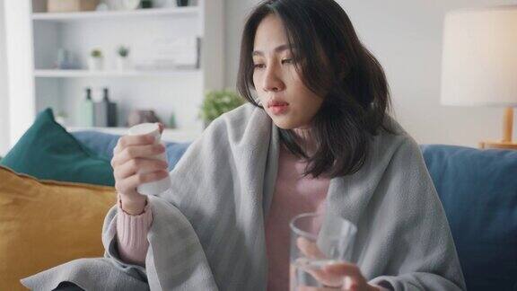 生病的亚洲妇女喝水喝处方药在家里客厅的沙发上盖着毯子忍受着头痛或抑郁