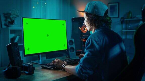 专业玩家在他强大的个人电脑上用五颜六色的霓虹灯玩在线视频游戏绿屏模拟年轻人戴着帽子客厅里有温暖的灯晚上