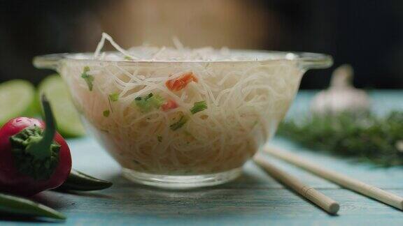 亚洲面条在玻璃碗与筷子和香料在蓝色的桌子上热汤加蒸汽