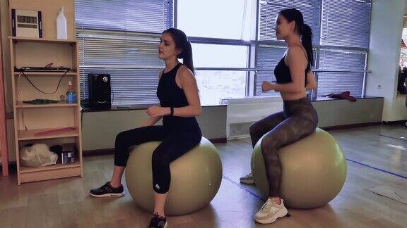 两个女孩在健身房里坐在健身球上跳跃