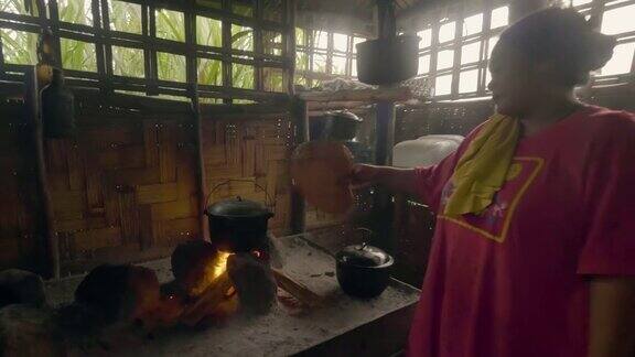 亚洲妇女吹火与风扇而烹饪食物在锅在竹屋篝火在亚洲的房子里传统的晚餐是在火锅里烧柴火