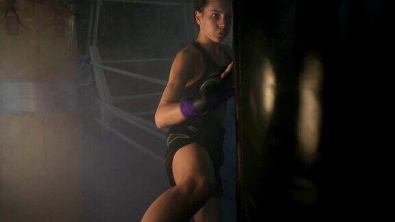 白种人强壮的女性自由搏击手运动与一个拳击袋在黑暗的健身房与烟和踢一个拳击袋与她的手和腿Slowmotion拍摄