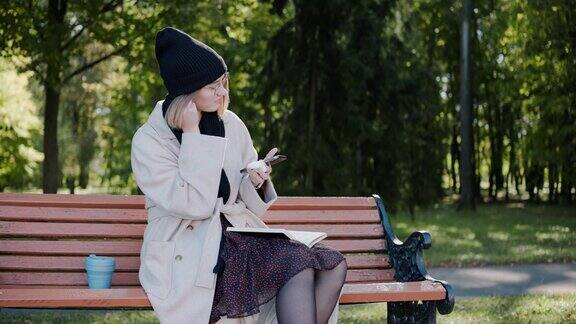 阳光明媚的秋天女孩坐在公园的长椅上戴着耳机微笑着