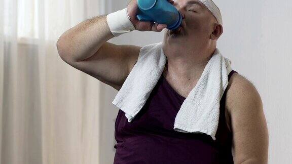 肥胖男性在运动后穿着运动服坐在垫子上喝着水