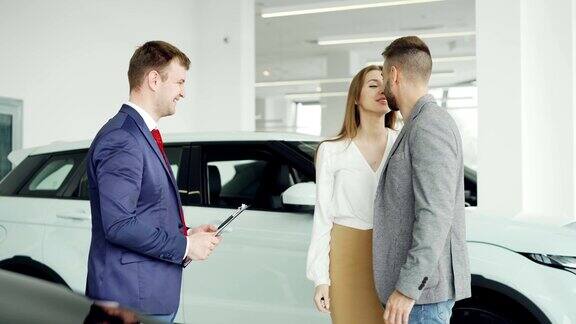 愉快的汽车经销商与年轻的买家英俊的男子握手然后给他的幸福的妻子钥匙链夫妇是拥抱和亲吻