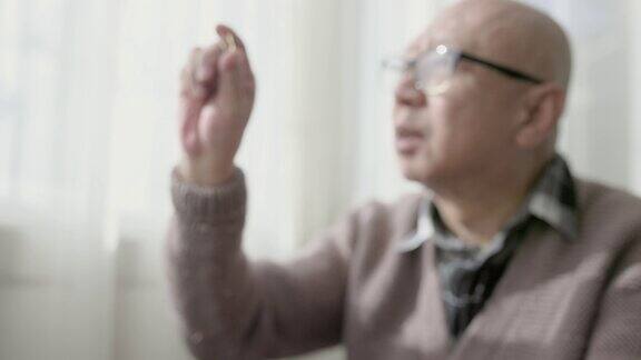 一位中国老人坐在窗边手里拿着一个胶囊戴着老花镜看电视