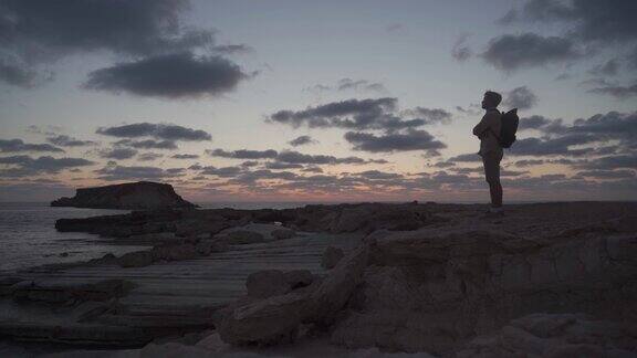 一名背着背包的男子在日落时分沿着塞浦路斯地中海附近的岩石海岸徒步旅行男游客们都很欣赏耶罗尼索斯岛或称圣岛它位于AgiosGeorgiosPegeias海岸附近