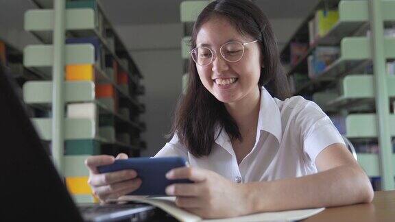 亚洲大学生女孩读书工作和网上购物与笔记本电脑和智能手机在公共图书馆做她的家庭作业