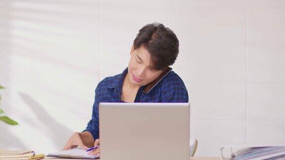 企业家英俊的商业亚洲男人穿着蓝色衬衫在笔记本电脑桌上工作接听手机与客户交谈以获得要求或订单微笑和快乐的工作在家里
