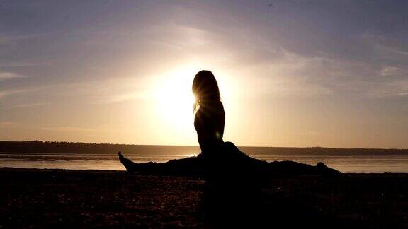 年轻女子在沙滩上做劈叉瑜伽姿势运动员美丽的剪影日落