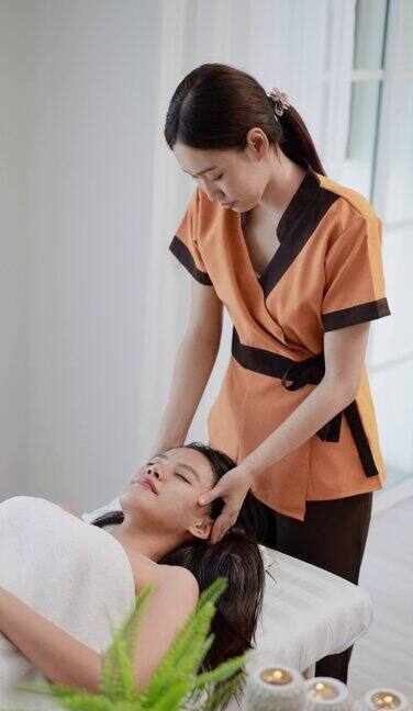 年轻的亚洲女人25岁在水疗沙龙做头部按摩女病人卧床闭眼按摩面部接受面部抗衰老治疗