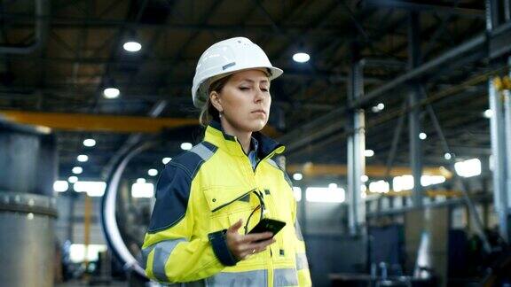 戴安全帽的女产业工人在穿过重工业制造厂时使用手机在各种金属制品项目的背景零件躺着