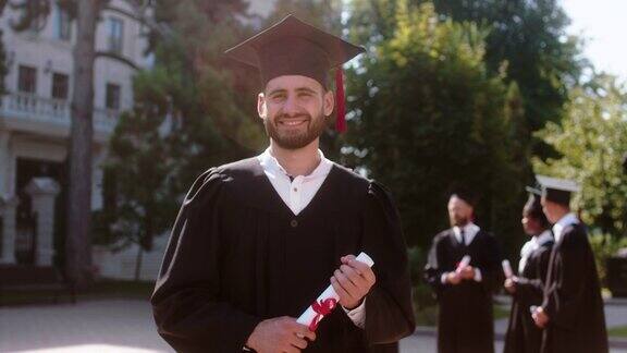 在一个阳光明媚的日子里在现代大学花园的毕业典礼上摆姿势的男毕业生他直视着镜头微笑着手里拿着毕业证书