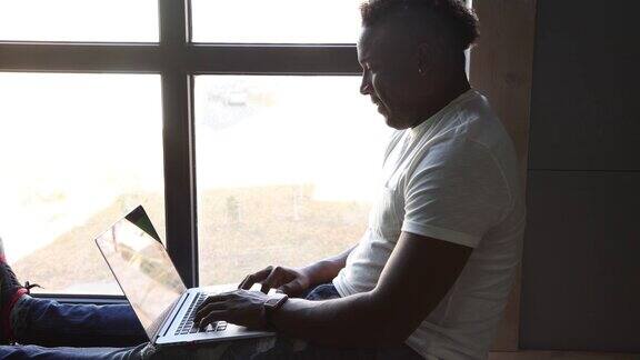 一个黑人坐在窗户边用笔记本电脑上网