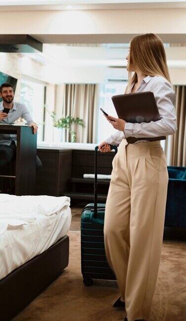 年轻的女商人带着手提电脑和行李箱离开酒店房间向同事打招呼商务青年离开酒店房间