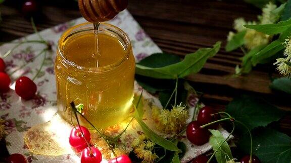打开玻璃罐液体蜂蜜和蜂蜜勺一束菩提花和红樱桃在木制表面射线的阳光黑暗的乡村风格