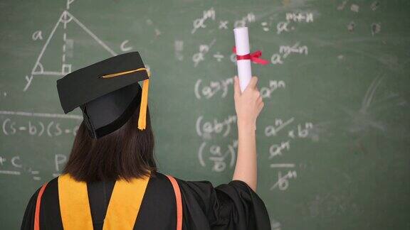 亚洲女毕业生身穿黑色长袍头戴金色流苏的黑色帽子站在教室的黑板前面