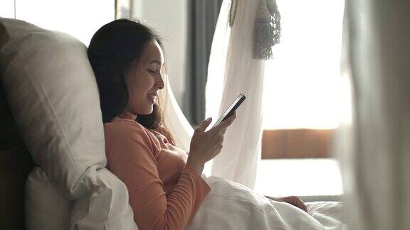 年轻的亚洲女子使用智能手机早上醒来后躺在床上