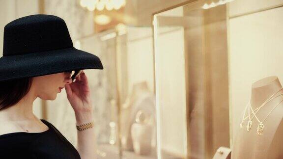 一位年轻漂亮的女士戴着帽子和太阳镜走到一家精品店的橱窗前兴致勃勃地看着商品奢华珠宝商店