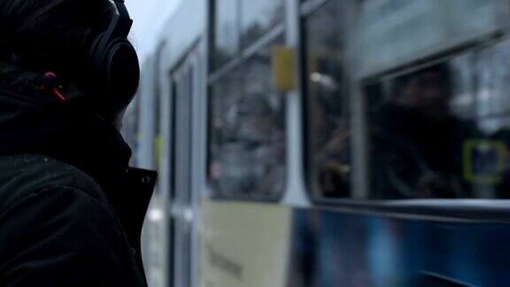 一个留着长头发留着胡子戴着耳机的年轻人在冬天站在一个电车站等电车的特写