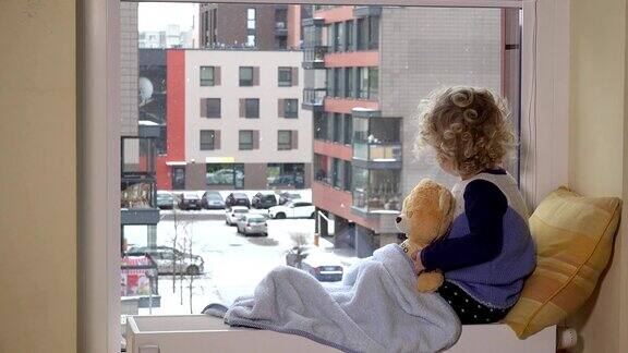 悲伤的蹒跚学步的男孩和最好的朋友泰迪熊看着雪花从窗户落下
