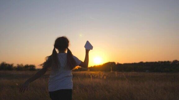 后视图可爱的小孩跑与纸飞机通过草地快乐的小女孩在草地上慢跑在夕阳背景下发射玩具飞机童年梦想