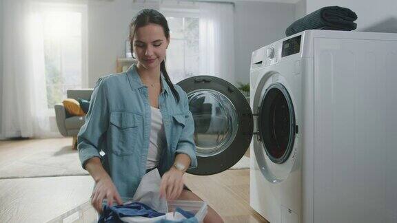 美丽微笑的黑发年轻女人坐在洗衣机前穿着普通的牛仔裤衣服她把脏衣服装在洗衣机里明亮宽敞的客厅现代的室内设计