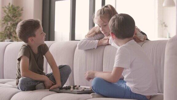 两个男孩坐在家里的沙发上下棋而姐姐在看他们