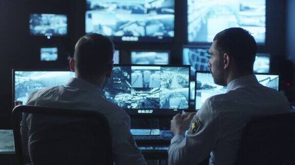 两名安全人员正在一间充满电脑显示屏的黑暗监控室中工作