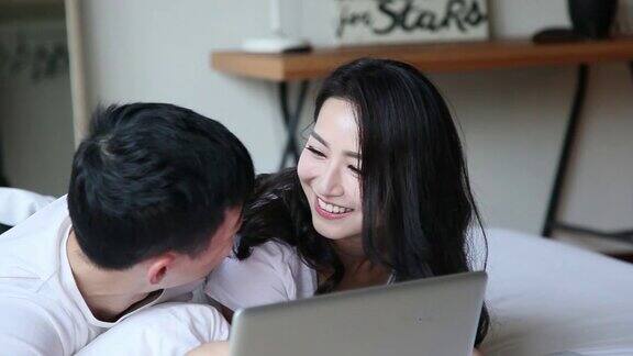亚洲华人夫妇拥抱用手提电脑微笑大笑早上在床上上网