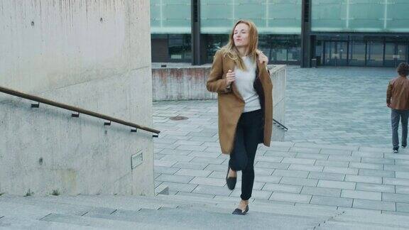 愉快和快乐的年轻女子积极跳舞而上行楼梯她穿着一件棕色长外套场景拍摄于商业中心旁边的城市混凝土公园天是光明的