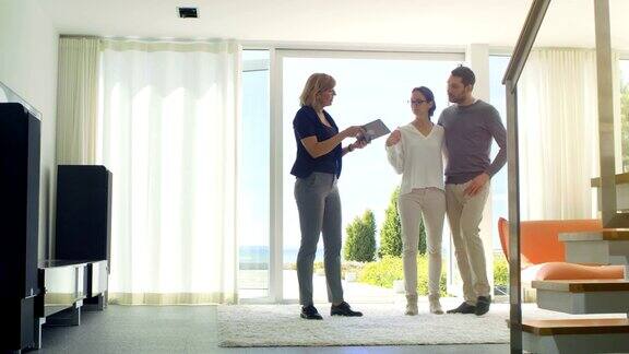 房地产经纪人向一对美丽的年轻夫妇展示用平板电脑控制的智能房子房子有落地窗和海景