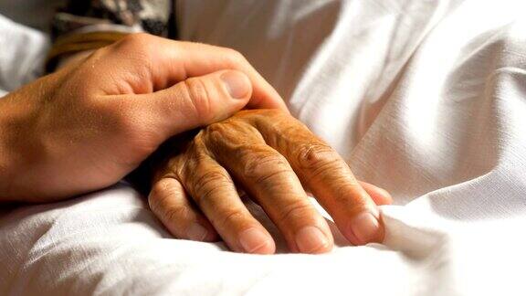 一个面目人非的男人轻轻地抚摸着生病的祖母的手给予支持儿子安慰着躺在医院病床上的老妈妈皱巴巴的手臂对年迈的父母表现出关爱的人