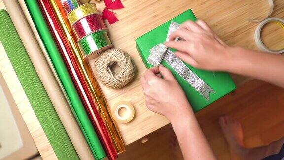 亚洲妇女的手在绿色的礼盒上用透明胶带系上银色的丝带圣诞节礼物在家里的桌子上用器皿、丝带、纸装饰在公寓的圣诞树和装饰品上俯瞰