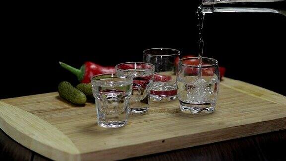 从一个瓶子里倒伏特加到三个玻璃杯里玻璃杯放在一块木板上上面放着胡椒、腌黄瓜和樱桃番茄