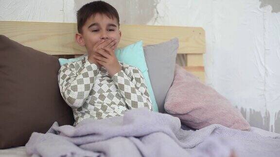 一个生病的小男孩坐在床上咳嗽并用毯子盖住自己高清