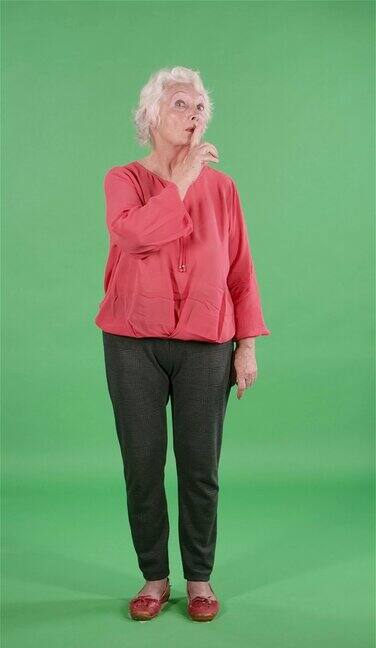 肖像老年妇女环顾四周做一个安静的手势秘密在演播室的绿色屏幕上灰头发的祖母穿着红色衬衫缓慢的运动垂直视频
