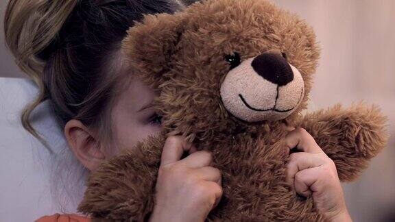 伤心的女孩躲在泰迪熊后面悲伤地看着摄像机领养程序