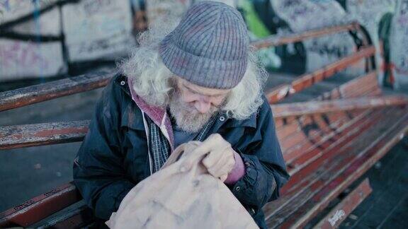 一个头发花白的流浪汉坐在屋外的长椅上在袋子里寻找食物