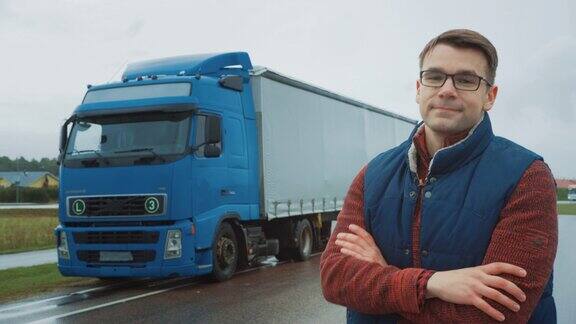 专业的年轻卡车司机交叉双臂和微笑在他身后停着一辆带货运拖车的蓝色长途半卡车