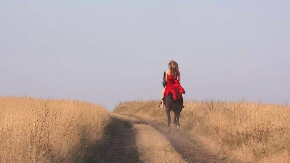 穿着红色长裙的年轻女孩骑着黑马在干燥的草原上