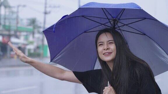 年轻的亚洲女子手持蓝色雨伞独自站在路边挥手呼叫或搭便车等待出租车或公共交通服务女孩的期待