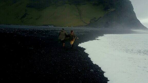 年轻的潮人夫妇在冰岛巨怪脚趾附近的黑色火山海滩上跑步的后视镜