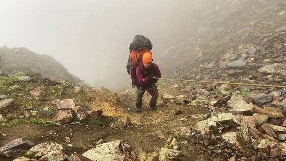 一名疲惫的登山者戴着橙色头盔背着一个大背包沿着小路爬上了山坡