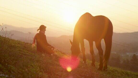 剪影:不认识的女人坐在她的种马旁边放牧在日出