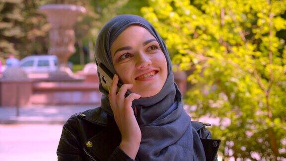 年轻漂亮的穆斯林女性的特写镜头戴着头巾在电话上愉快地微笑在城市的街道上的城市户外