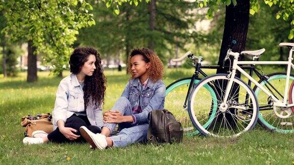 一位漂亮的非裔美国妇女和她的欧洲朋友坐在公园的草地上聊天女孩们有说有笑自行车和树在背景阳光灿烂的一天